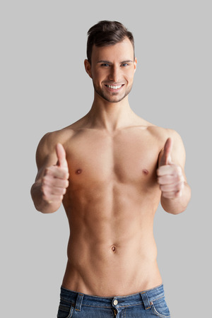 Join a healthy lifestyle. Cheerful young muscular man looking at camera and gesturing while standing isolated on grey backgroundの写真素材