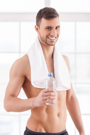 Rehydrating after workout. Handsome young muscular man with towel on shoulders holding a bottle with water and smilingの写真素材