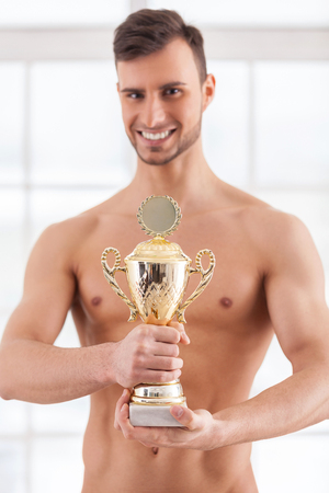 Real achievement. Handsome young muscular man holding a golden trophy in his hands and smilingの写真素材