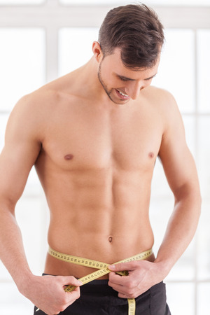 Keeping body in perfect shape. Young muscular man measuring his waist with measuring tape and smilingの写真素材