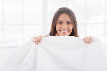 Good morning! Cheerful young woman sitting in bed and looking out of the blanketの写真素材