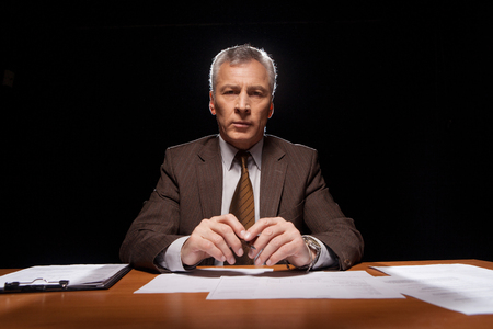 Serious senior man in formal wear sitting at his working place and looking at camera while isolated on black background の写真素材
