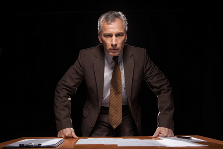 Serious senior man in formal wear looking at camera while leaning at the table and isolated on blackの写真素材