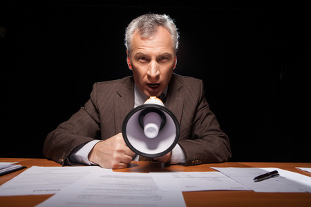 Furious senior man in formal wear sitting at his working place and shouting at megaphone while isolated on black background の写真素材
