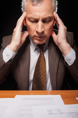Senior man in formal wear sitting at his working place and holding head in hands while isolated on black background の写真素材