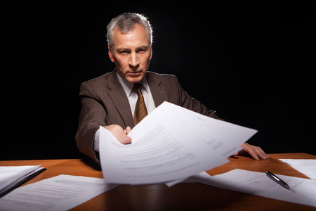 Serious senior man in formal wear sitting at his working place and stretching out documents while isolated on black background の写真素材