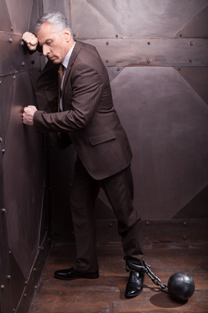 grey hair senior man in formal wear with the shackles on his leg leaning at the wallの写真素材