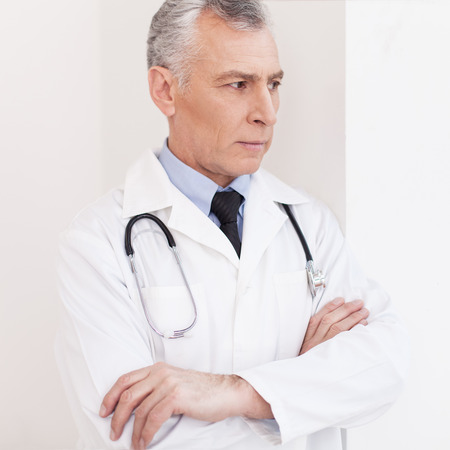 Senior grey hair doctor in uniform looking away and keeping hands claspedの写真素材