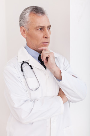 Senior grey hair doctor in uniform looking away and holding hand on chinの写真素材