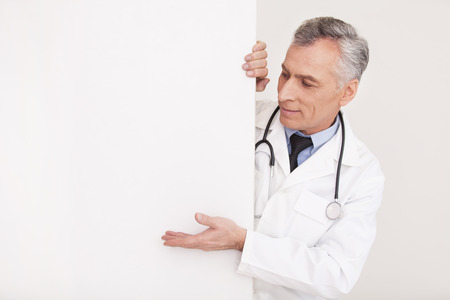 Senior grey hair doctor in uniform looking out of copy space and pointing it while isolated on whiteの写真素材
