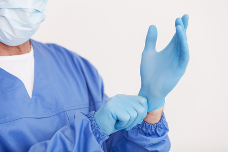 doctor in surgical mask adjusting his protective glove while standing isolated on whiteの写真素材