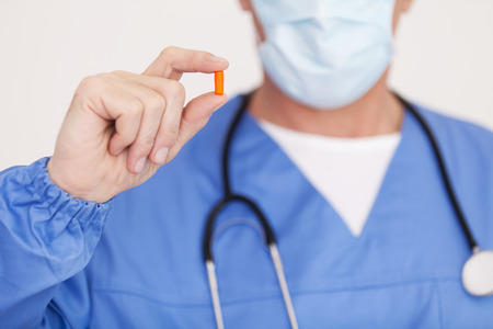 doctor in surgical mask showing a pill while standing isolated on whiteの写真素材