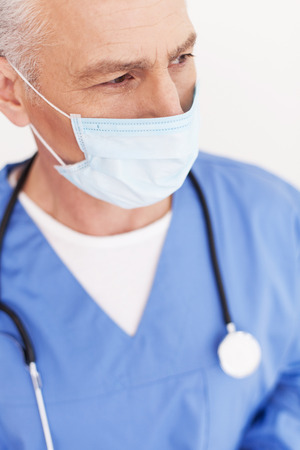 thoughtful senior doctor in surgical mask looking away while standing isolated on whiteの写真素材