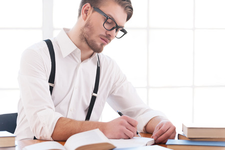 Author at work. Confident young man in shirt and suspenders writing something in note pad while sitting at his working placeの写真素材