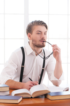 Waiting for inspiration. Thoughtful young man in shirt and suspenders holding glasses and looking away while sitting at his working placeの写真素材