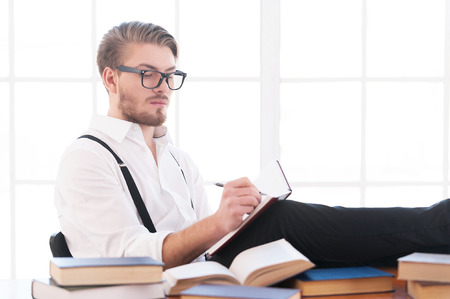 Writer at work. Handsome young man in shirt and suspenders writing something in notebook while sitting at his working placeの写真素材