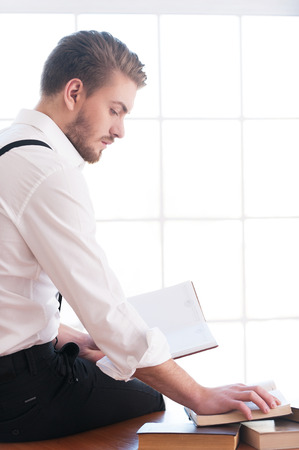 Handsome bookworm. Rear view of thoughtful young man in shirt and suspenders sitting on the table and reading a book laying on itの写真素材