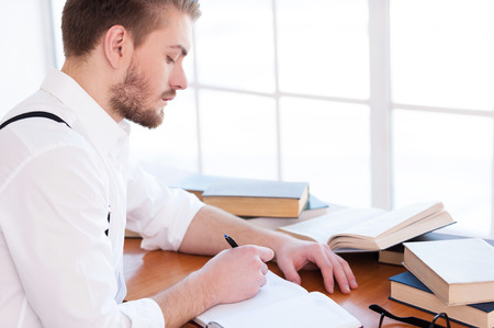 Confident and inspired. Side view of confident young man in shirt and suspenders writing something in note pad while sitting at the table with books laying on itの写真素材