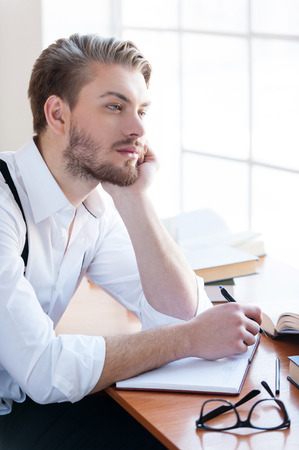 Waiting for inspiration. Handsome young man in shirt and suspenders holding head in hand and looking away while sitting at the table with books laying on itの写真素材