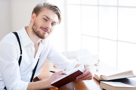 Inspired author. Handsome young man in shirt and suspenders writing something in note pad and smiling at camera while sitting at his working placeの写真素材