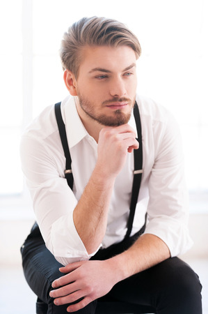 Looking for inspiration. Thoughtful young man in shirt and suspenders holding hand on chin and looking away while sitting on the chairの写真素材