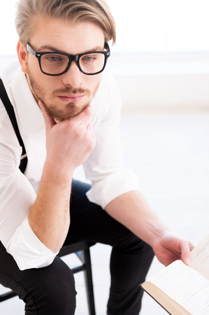 What an interesting fact! Top view of thoughtful young man in shirt and suspenders holding a book and looking away while sitting on the chairの写真素材