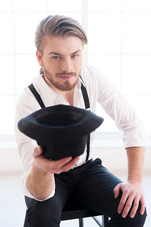 In style. Handsome young man in shirt and suspenders stretching out hand with hat in it and looking at camera while sitting on the chairの写真素材