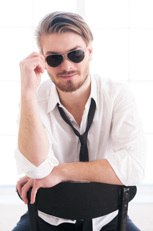 Feeling flirty. Thoughtful young man adjusting his sunglasses and looking at camera while sitting on the chairの写真素材