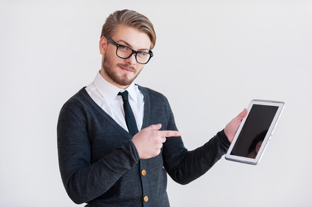 It will make your life easier. Handsome young man in glasses holding a digital tablet and pointing it while standing against grey backgroundの写真素材