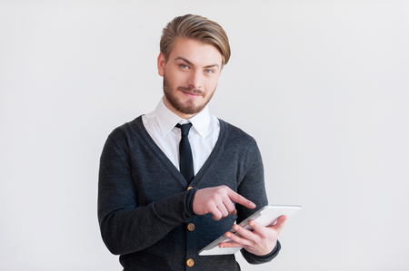 It will make your life easier. Handsome young man in glasses holding a digital tablet and smiling while standing against grey backgroundの写真素材