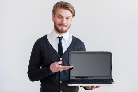 Your advertising here! Handsome young man holding a laptop and pointing it while standing against grey backgroundの写真素材