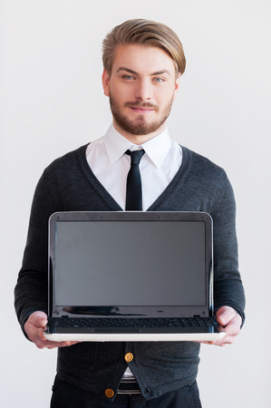 Copy space on monitor. Handsome young man holding a laptop and smiling while standing against grey backgroundの写真素材