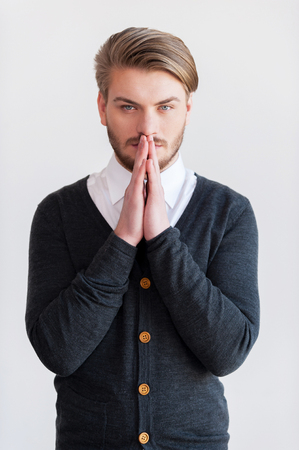 Man praying. Handsome young beard man keeping hands clasped and looking at camera while standing against grey backgroundの写真素材