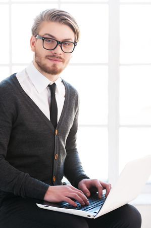 Handsome with laptop. Handsome young man working on laptop and smiling at camera while sitting on the chairの写真素材