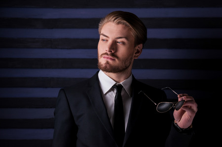 Confident and good looking. Portrait of handsome young man in formalwear holding sunglasses and looking away while standing against striped backgroundの写真素材
