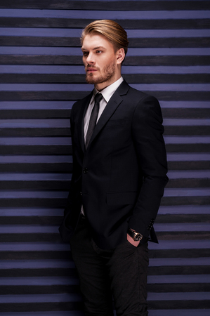 Thoughtful handsome. Side view of handsome young man in formalwear holding hands in pockets and looking away while standing against striped backgroundの写真素材