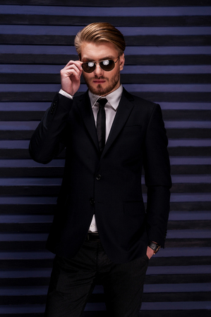 Confident in his handsome look. Handsome young man in formalwear adjusting his sunglasses and holding one hand in pockets while standing against striped backgroundの写真素材