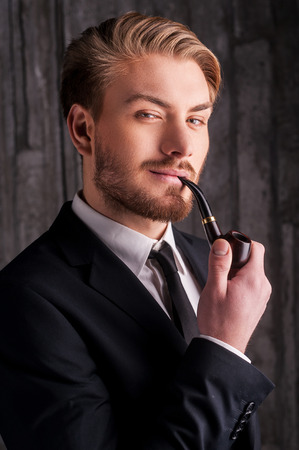 Elegance and masculinity. Portrait of handsome young man in formalwear smoking a pipe and smiling at camera の写真素材
