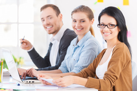 Coming up with fresh design solutions. Three cheerful young colleagues sitting together at their working place and smiling at cameraの写真素材
