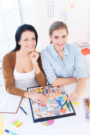 Creative executive. Top view of two young beautiful women smiling at camera while sitting at their working placeの写真素材