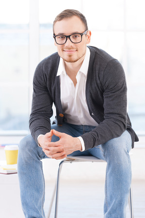 Looking for inspiration. Thoughtful young man in shirt looking at camera and smiling while sitting on the chairの写真素材