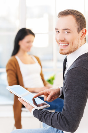Creative team  Cheerful young man in glasses using digital tablet and smiling while his colleagues working on the backgroundの写真素材