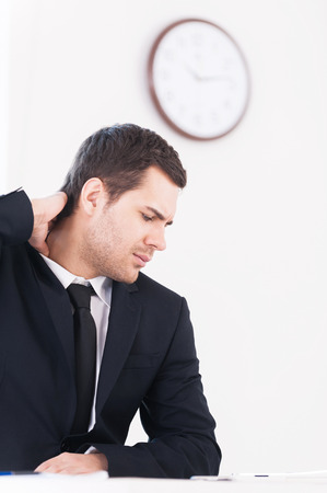 No time for break. Depressed young man in formalwear looking away and touching his neck while sitting at his working place with wall clock on backgroundの写真素材