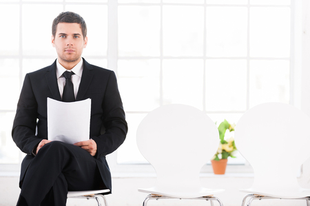 Waiting for job interview. Confident young man in formalwear sitting at the chair and holding head in hand while other chairs standing in a rowの写真素材