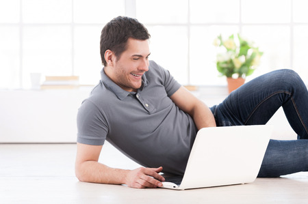 Surfing the net at home. Cheerful young man lying on the floor and using computerの写真素材