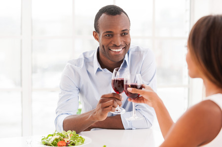 Dinning together. Beautiful young African couple sitting together at the table and holding wineglassesの写真素材