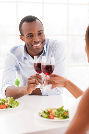 Having a dinner together. Cheerful young African couple sitting together at the table and holding wineglassesの写真素材