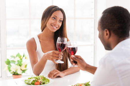 Good wine for special date. Beautiful young African couple sitting together at the table and holding wineglassesの写真素材