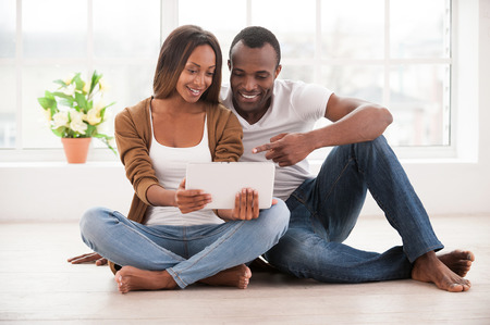 Working at their new tablet. Beautiful young African couple using digital tablet while sitting together on the floor at their apartments の写真素材