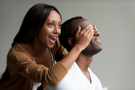 I have a surprise for you! Beautiful young African woman covering her boyfriend eyes with hands and smiling while isolated on grey background の写真素材